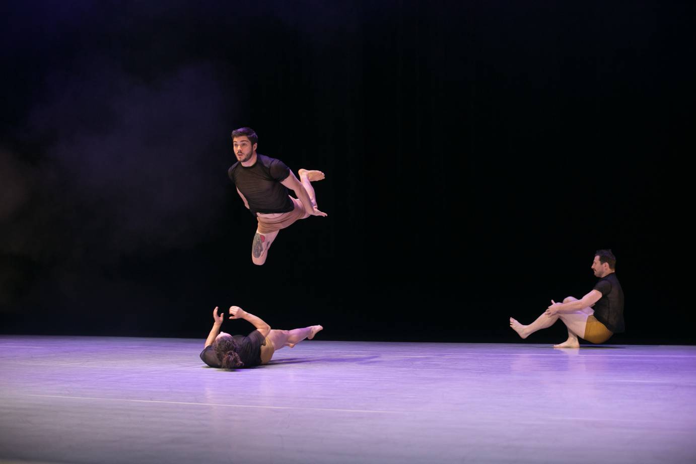 Two dancers roll on the floor while another jumps in the air parallel above one of them.